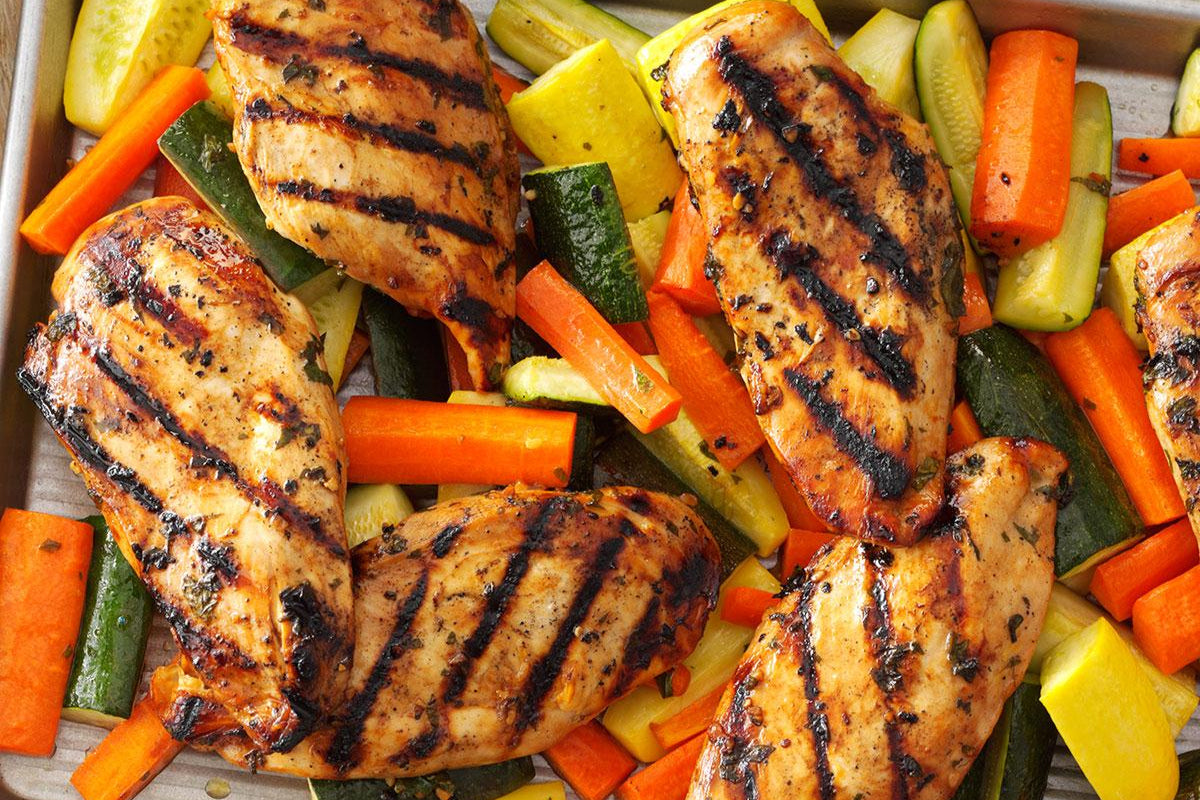 A dish of grilled chicken with mixed vegetables, including carrots, zucchini, broccoli, and cauliflower, served in a metal pan.